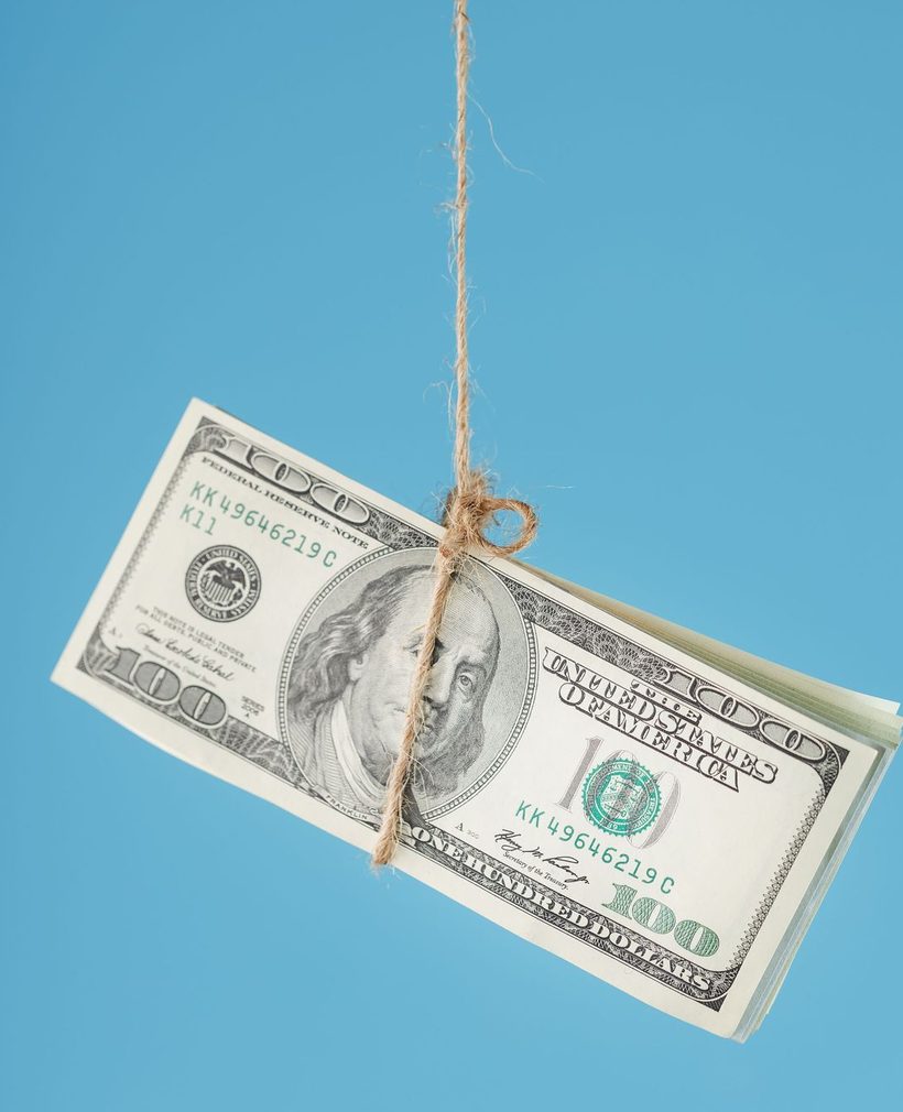 A bundle of one hundred dollar bills tied with twine, hanging against a blue background